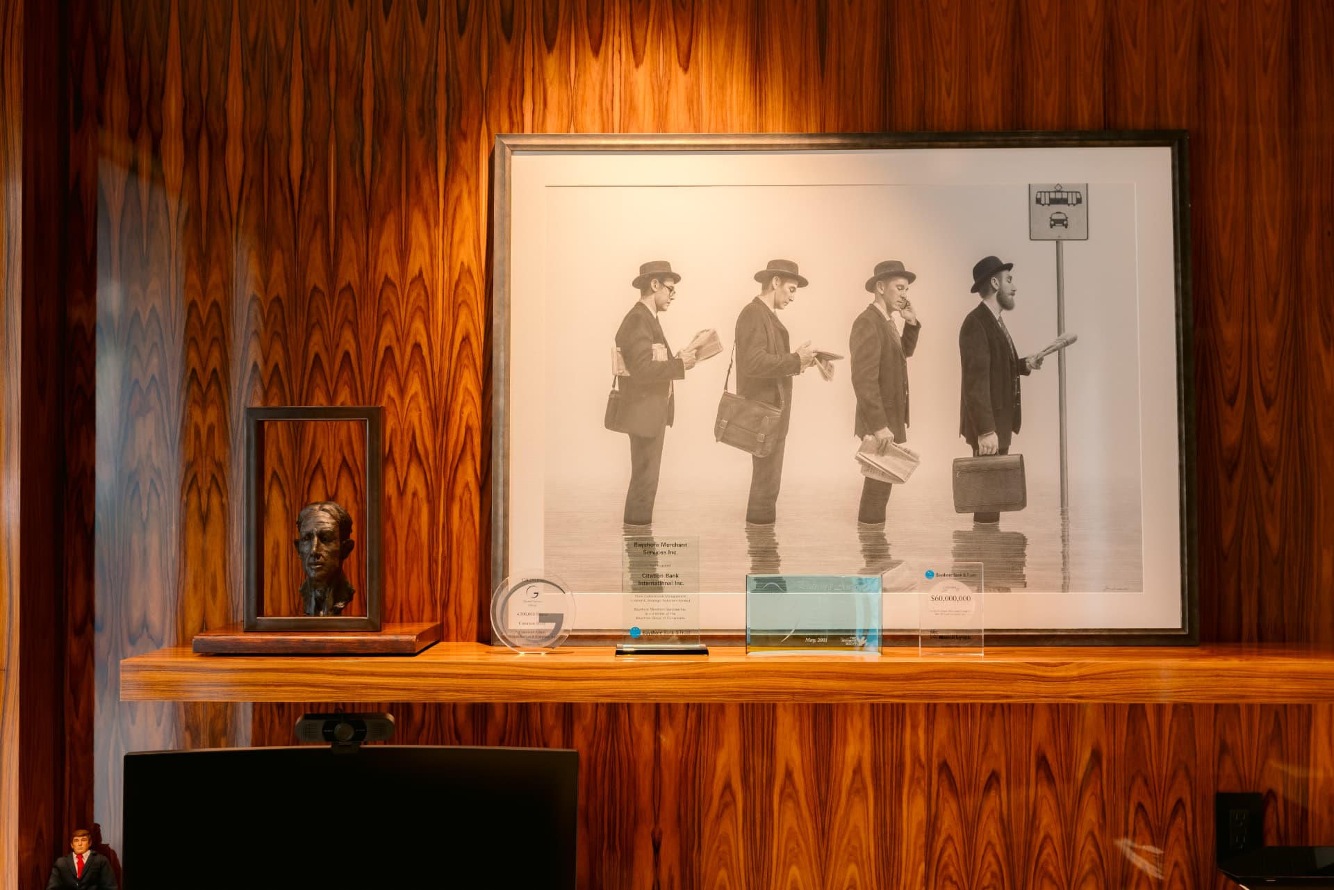 A framed print of four men waiting at a transit stop, hung above a rosewood credenza with a bronze bust and awards